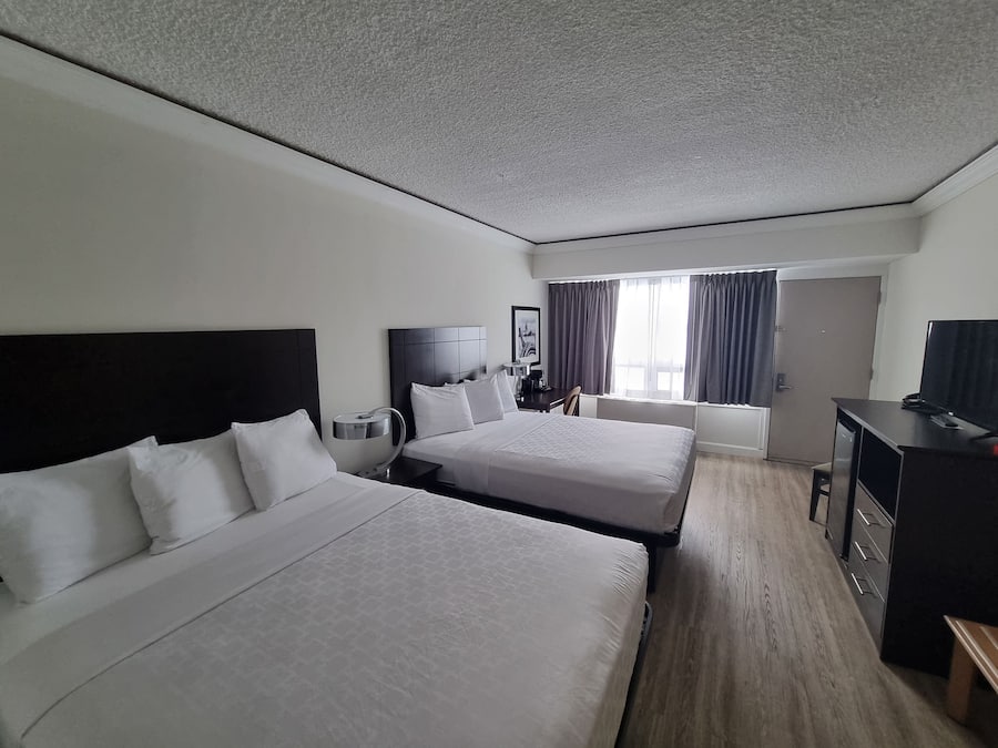 Quarto de hotel com duas camas, uma escrivaninha, uma cadeira, uma televisão e uma janela com cortinas.