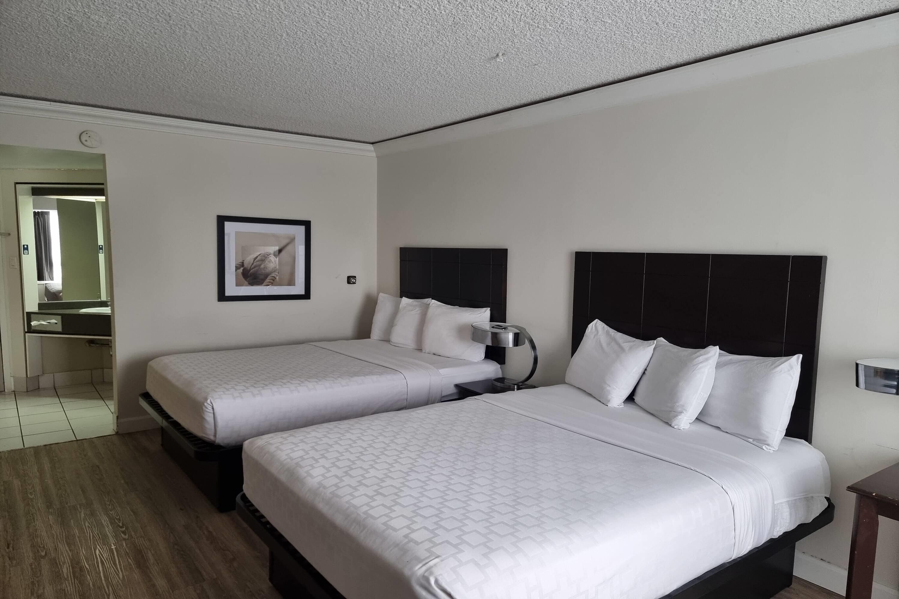 Habitación de hotel con dos camas, una mesita de noche con lámpara, un cuadro enmarcado en la pared y un baño visible a través de una puerta abierta.