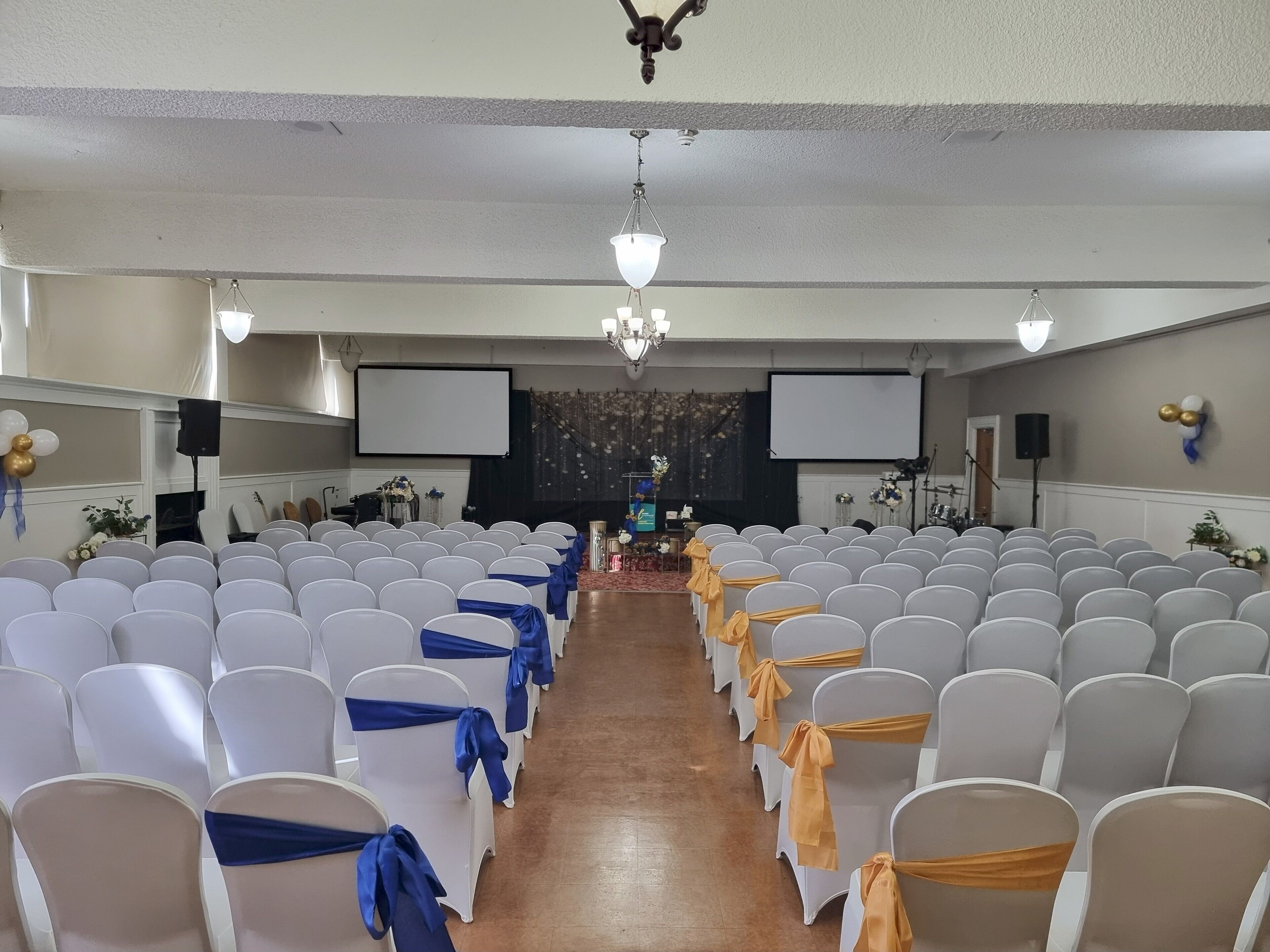 Un salón de conferencias con filas de sillas blancas, algunas con cintas azules y amarillas, un escenario con un atril y dos pantallas grandes.