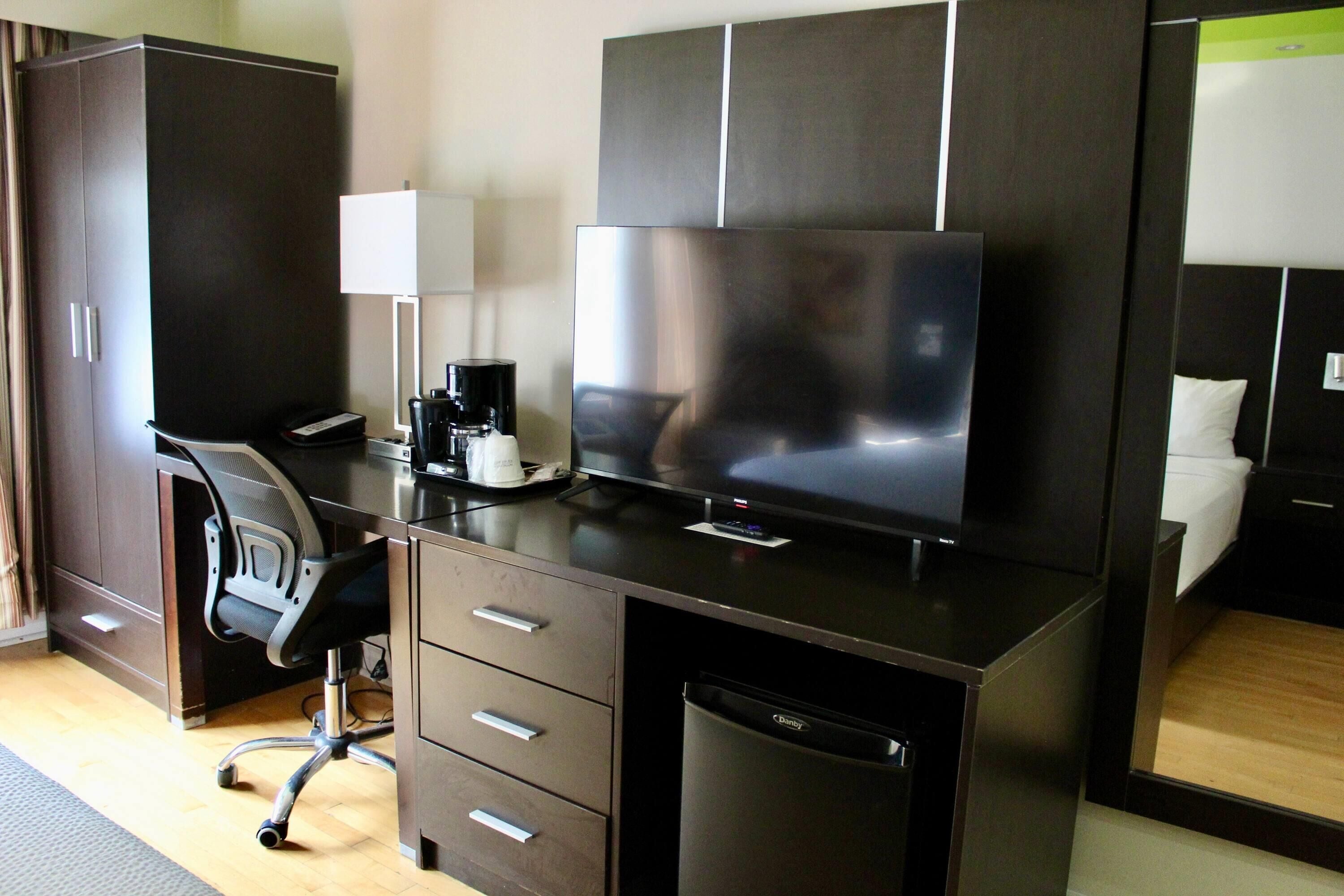 A modern hotel room with a dark wood desk, a flat-screen TV, a coffee maker, a chair, and a built-in dishwasher.