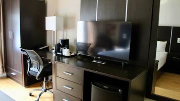 A modern hotel room with a dark wood desk, a flat-screen TV, a coffee maker, a chair, and a built-in dishwasher.