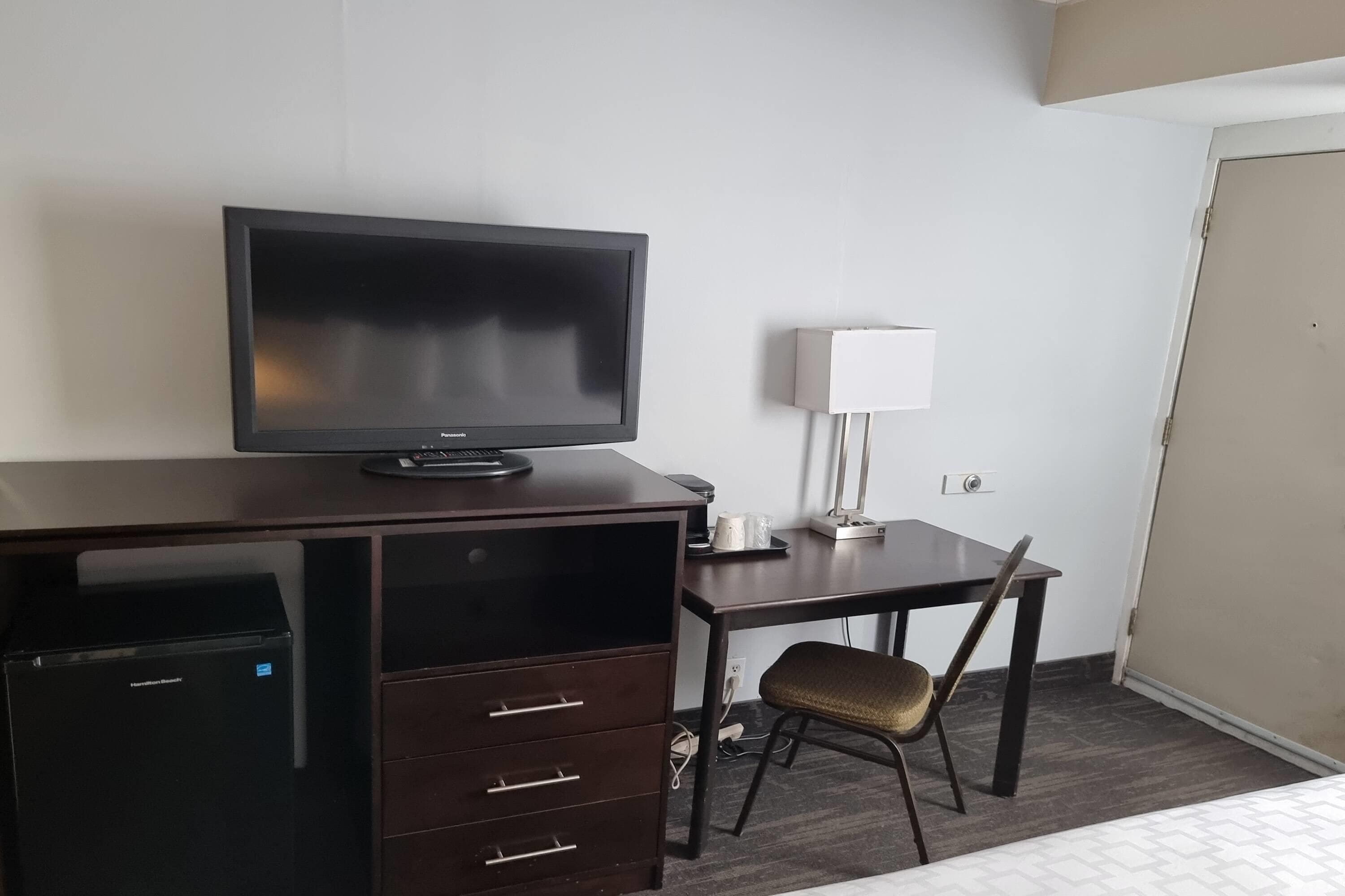 A hotel room with a dark wooden desk, a television, a lamp, a chair, and a minibar.