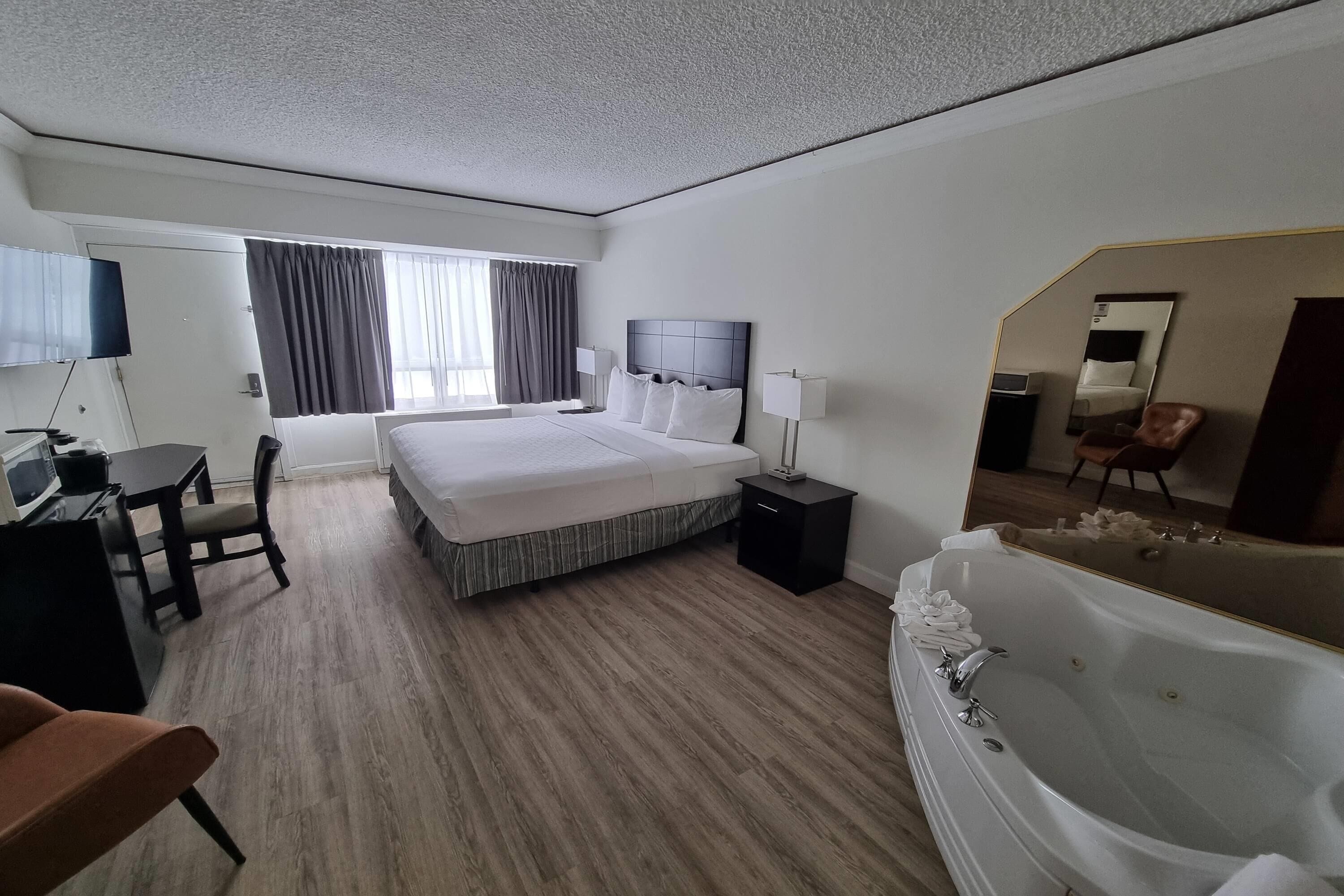 A hotel room with a large bed, a desk with a chair, a desk lamp, a TV, a large mirror, and a bathtub.