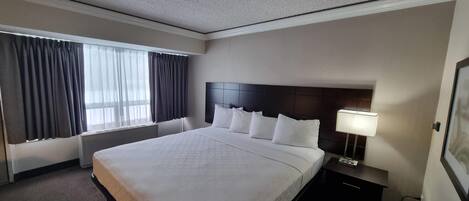 Ein Hotelzimmer mit einem großen Bett, einem Nachttisch mit Lampe, einem Fenster mit Vorhängen und einem an der Wand montierten Fernseher.