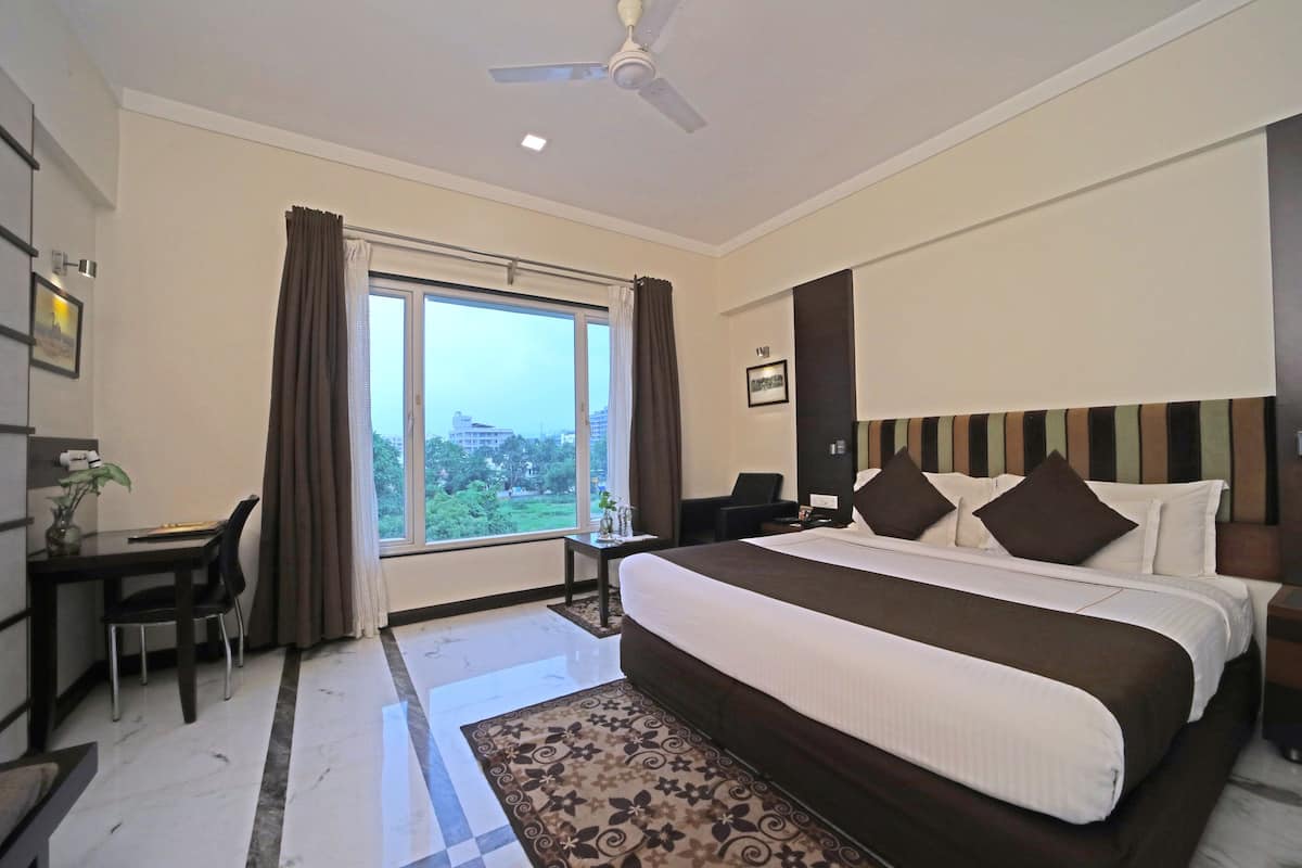A hotel room with a large bed, a desk with a chair, a small table, a nightstand, and a view of the outdoors.
