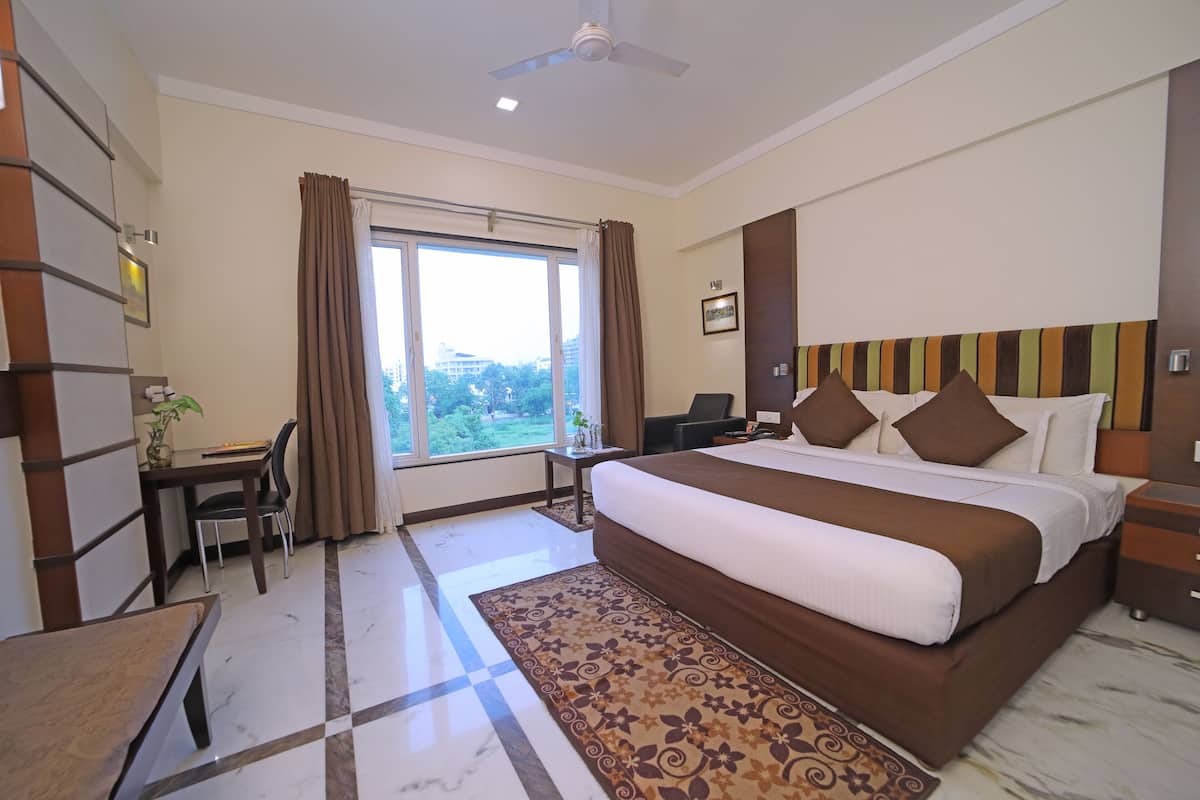 A hotel room with a large bed, a desk, and a chair. There is a window with curtains, a ceiling fan, and a view of greenery outside.