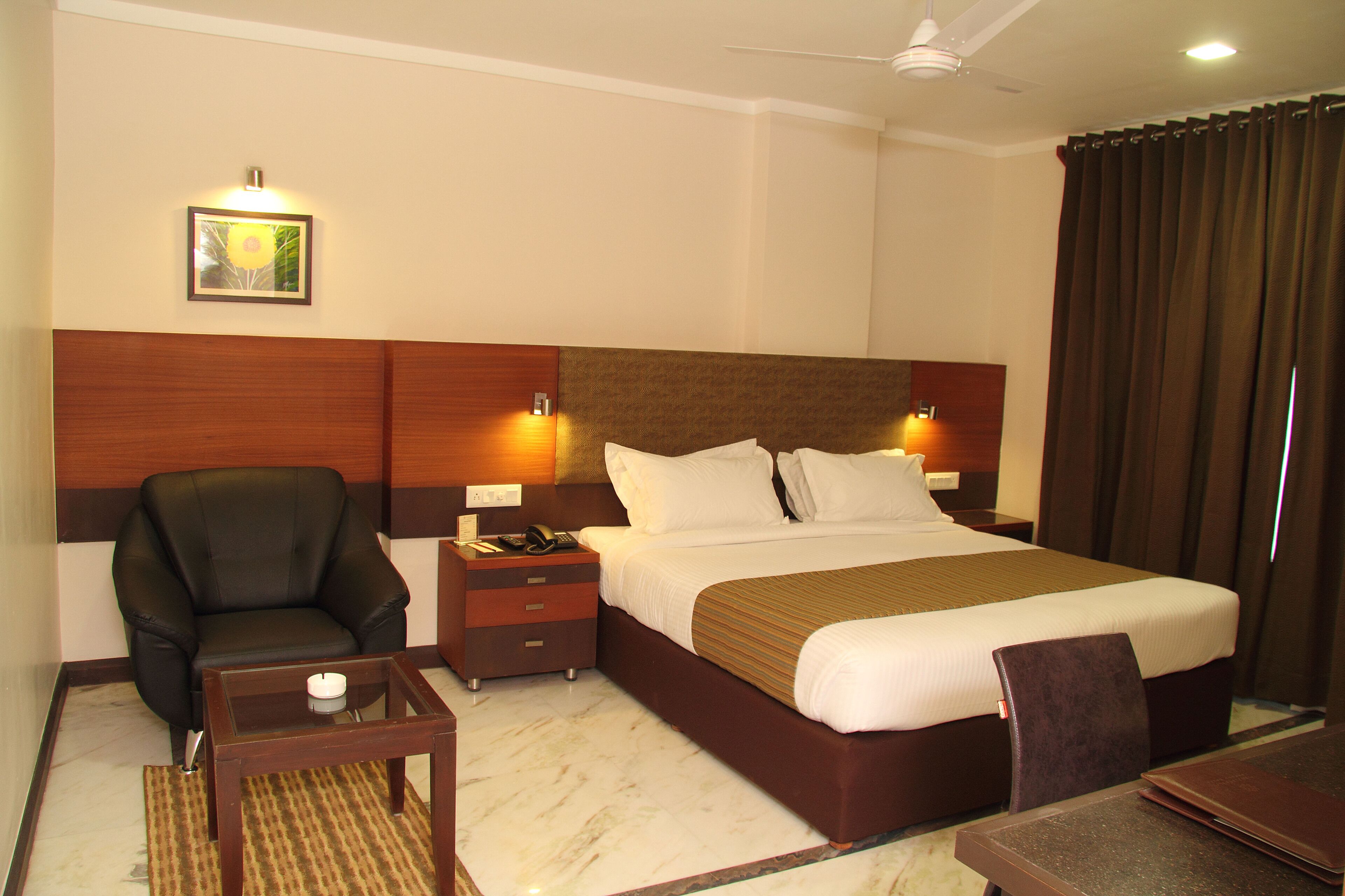 A hotel room with a large bed, a chair, a small table, a nightstand, and a ceiling fan.
