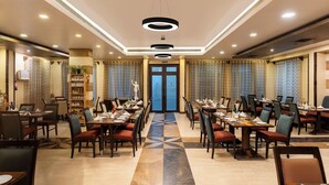 A restaurant with multiple dining tables and chairs, a central aisle, and a patterned floor. - Amantra Comfort Hotel (Udaipur)