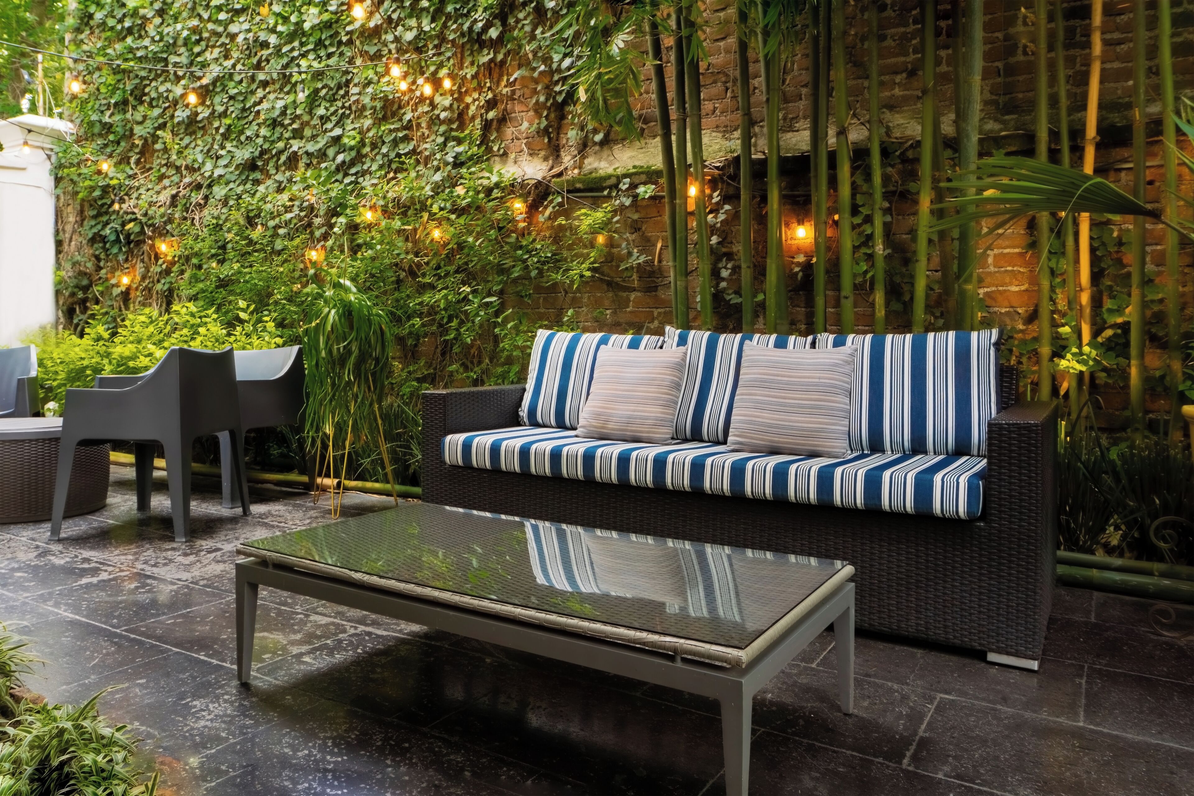 Un espace extérieur aménagé avec un canapé et des fauteuils, une table à plateau en verre et un mur de briques agrémenté de plantes et de guirlandes lumineuses.