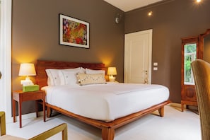 A hotel room with a large bed, two bedside lamps, a framed artwork on the wall, a door, and a window with a view of greenery.
