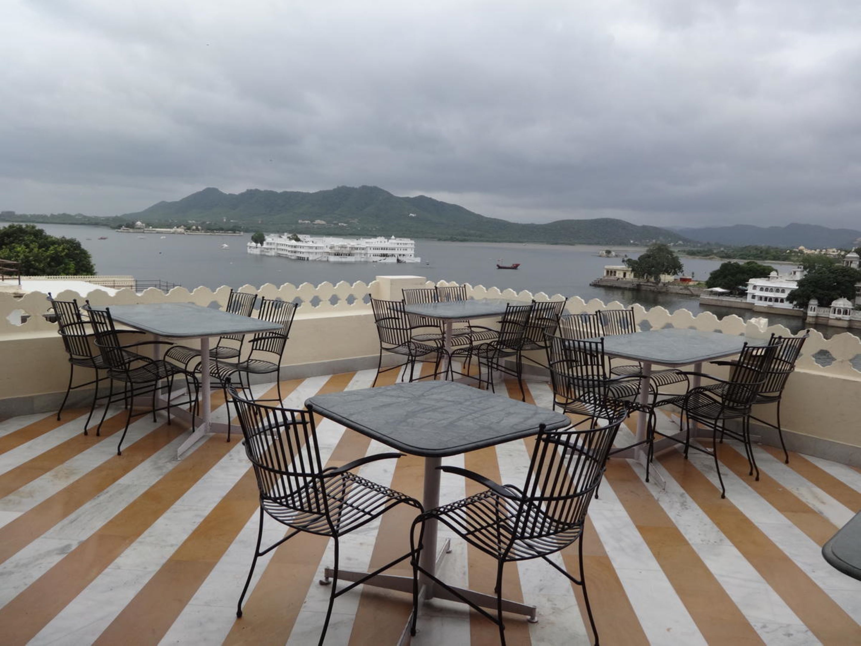 Una terraza con mesas y sillas con vista a un lago con barcos y una cadena montañosa distante.