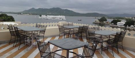 Una terraza con mesas y sillas con vista a un lago con barcos y una cadena montañosa distante.