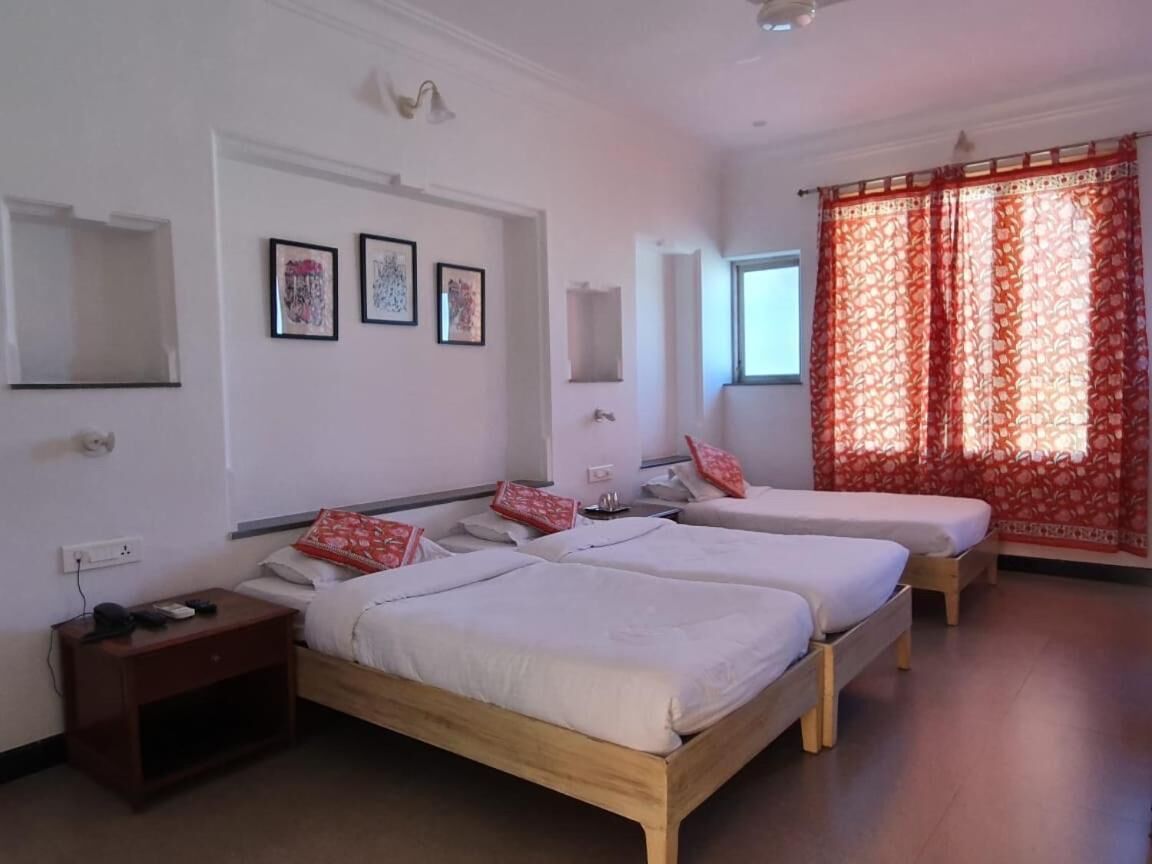 A hotel room with two single beds, a nightstand, a small table, and a window with red curtains.