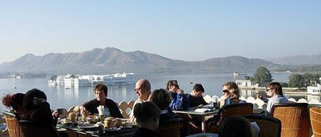 Un grupo de personas cenando en una mesa al aire libre con vista a montañas y un cuerpo de agua.