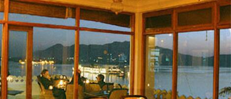 A dining area with tables and chairs, large windows with a view of a body of water and distant mountains, and hanging light fixtures.