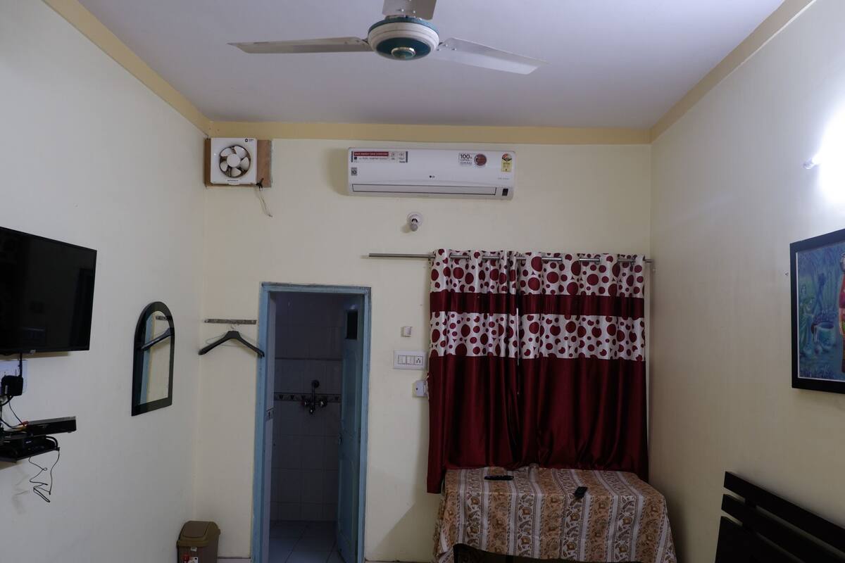 A bedroom with a bed, a ceiling fan, an air conditioner, a TV, a door, and a small trash bin.