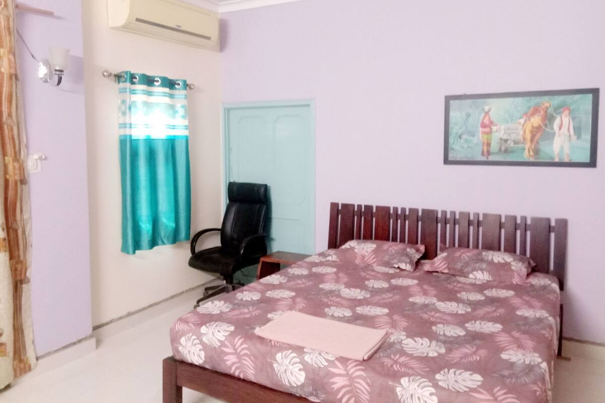 A bedroom with a bed, a chair, an air conditioning unit, a door, and a wall-mounted painting.