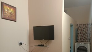 Una habitación pequeña con un televisor montado en la pared, una mesita de madera con un teléfono y controles remotos, y piso de baldosas.