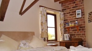 A bedroom with a bed, a window with curtains, a brick wall, a wooden shelf with items, and a framed picture. - Bluebell Farm (Worcester)