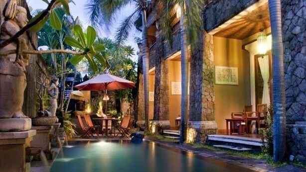 Un edificio de piedra con piscina, mobiliario exterior y plantas tropicales.
