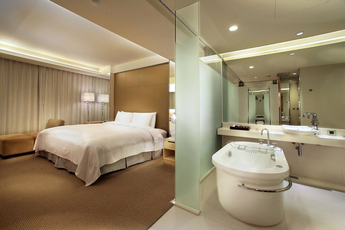 A hotel room with a bed, bedside table, lamp, and a large mirror above the sink.