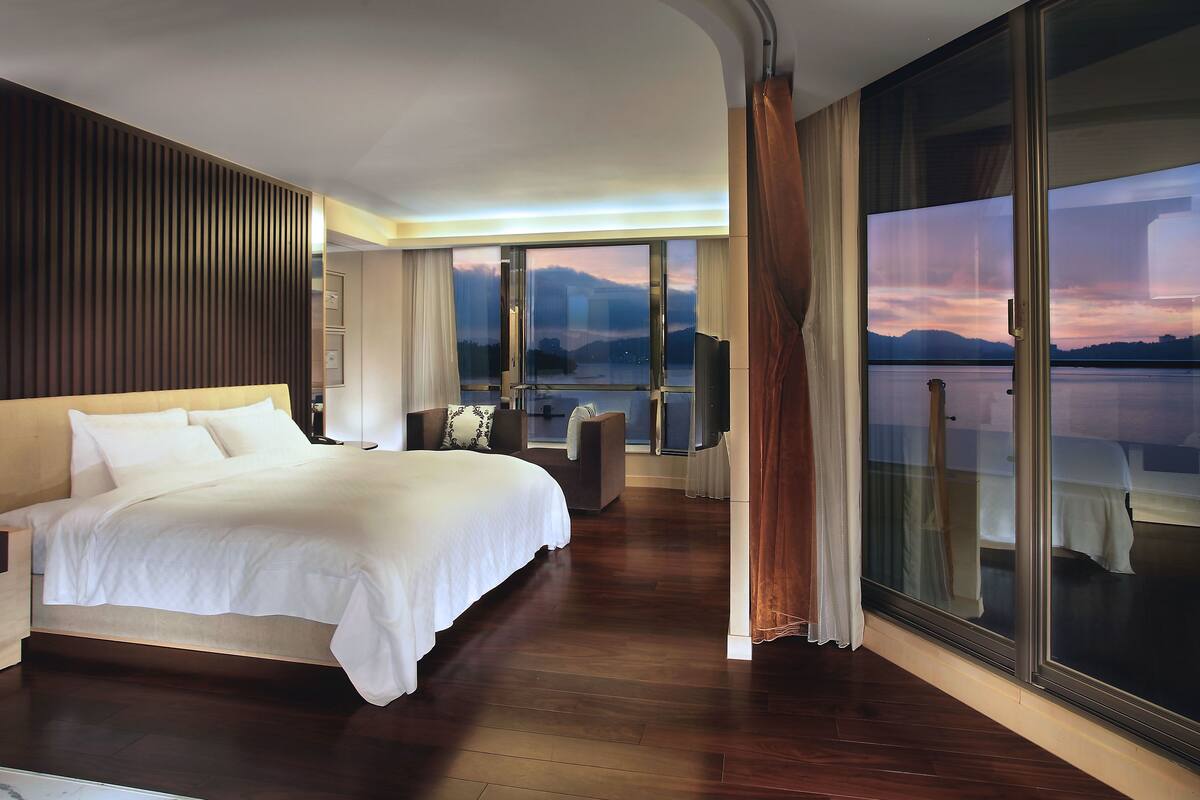 A modern hotel room with a large bed, a seating area, and a view of a body of water and mountains.