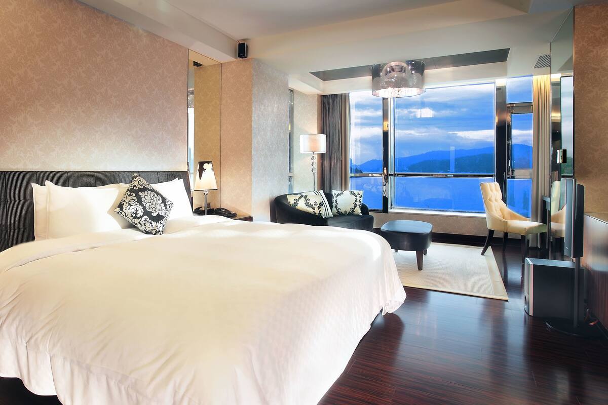 A large bed with white linens, a nightstand with a lamp, a sofa with patterned cushions, a chair, and a television.