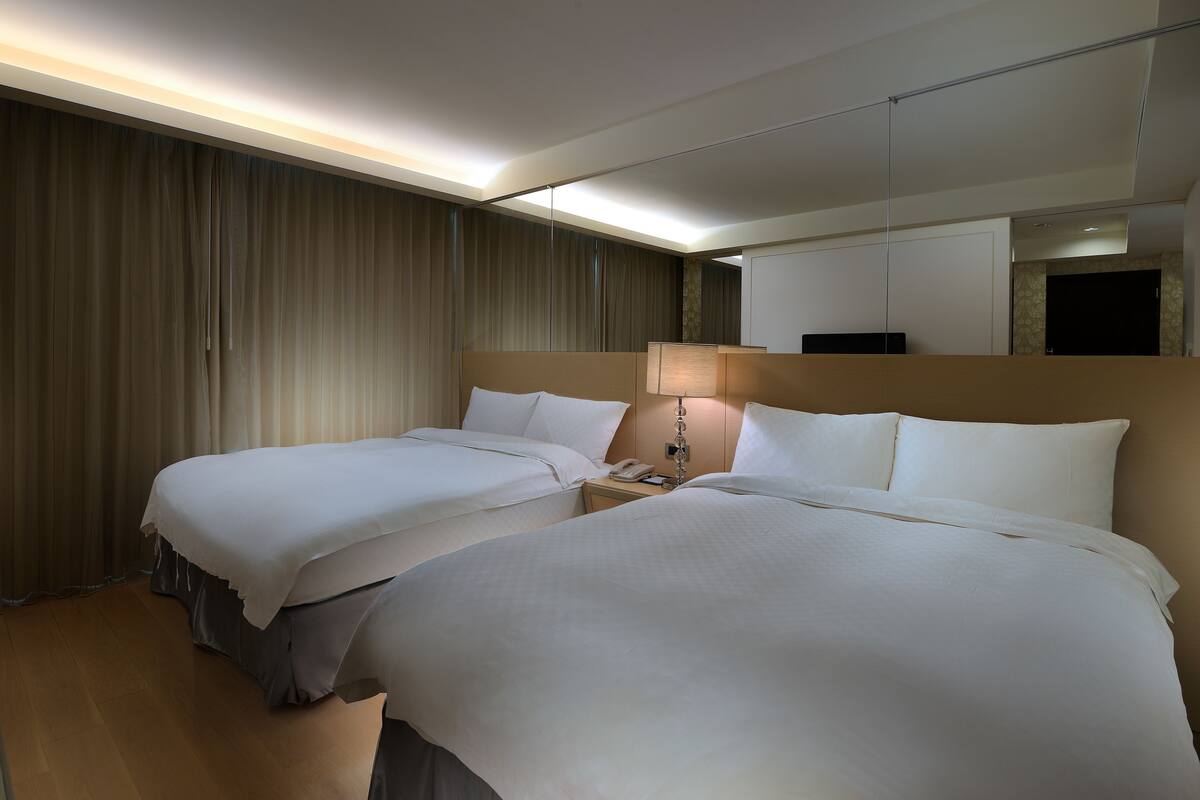 A hotel room with two beds, a nightstand with a lamp, and a large mirror.