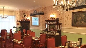 A dining area with red chairs, tables set with white plates and silverware, a chandelier, and a wall clock. - Pelham Lodge (Blackpool)