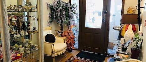 Una habitación con un aparador de vidrio lleno de diversos objetos, una silla, una planta grande en maceta y una alfombra estampada.
