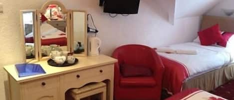 Habitación de hotel con cama, una silla roja, cómoda con cajones, espejo, televisor de pantalla plana montado en la pared y una mesita de noche con taza y platillo.