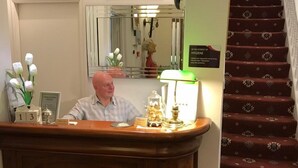 A hotel reception desk with a wooden counter, a clock, a flower vase, and a sign indicating hygiene interest. - Pelham Lodge (Blackpool)