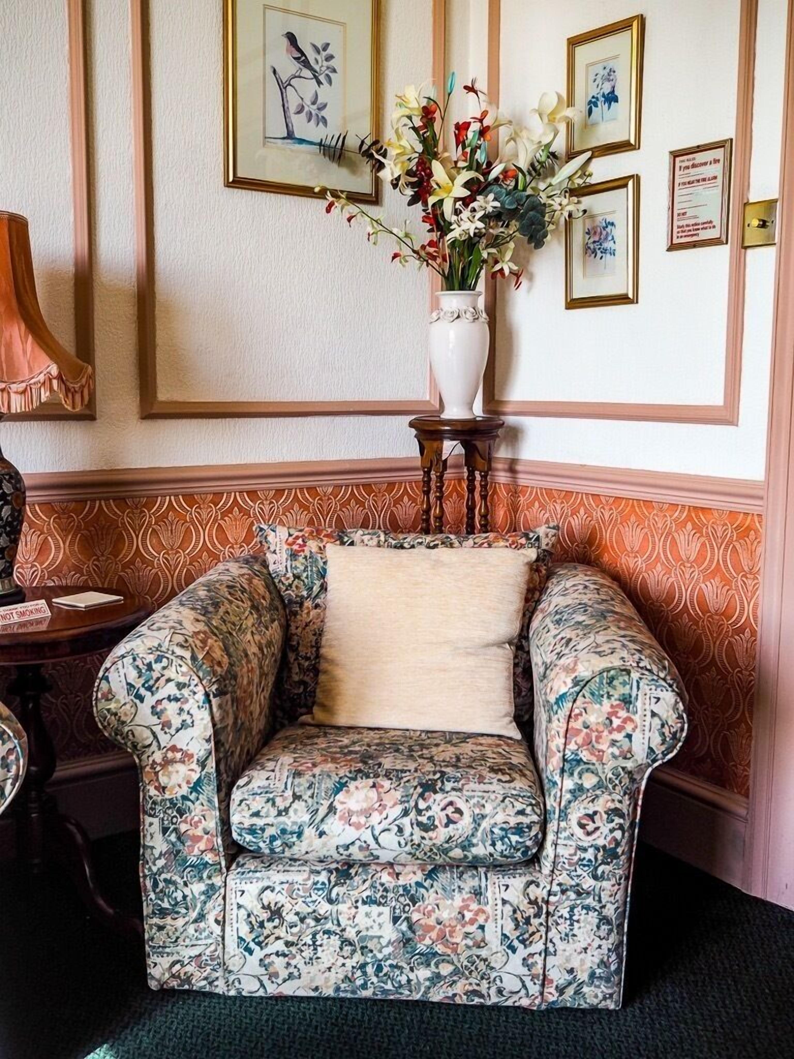 Una habitación de esquina con un sillón estampado con motivos florales, un jarrón con flores y cuadros en la pared.