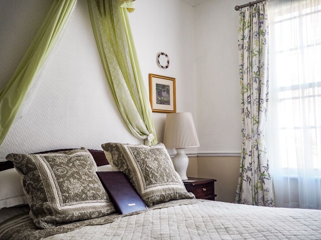 Une chambre à coucher avec un lit, des rideaux verts, une lampe de chevet, un tableau encadré au mur et une fenêtre ornée de rideaux à motifs floraux.