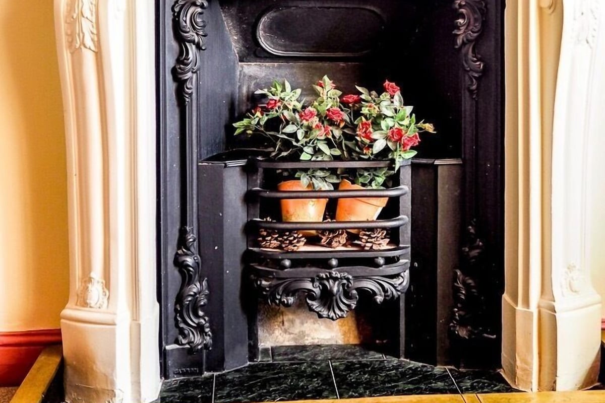 Une cheminée ornée d’un manteau de couleur blanche et agrémentée de motifs décoratifs, avec des touches noires, flanquée de deux plantes en pot aux fleurs rouges.