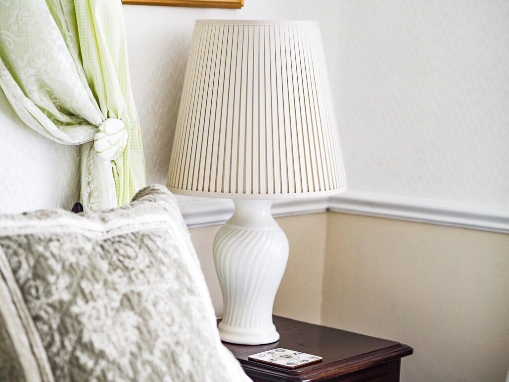 A white lamp on a wooden nightstand, a patterned throw pillow, and a green curtain tied back on a wall.