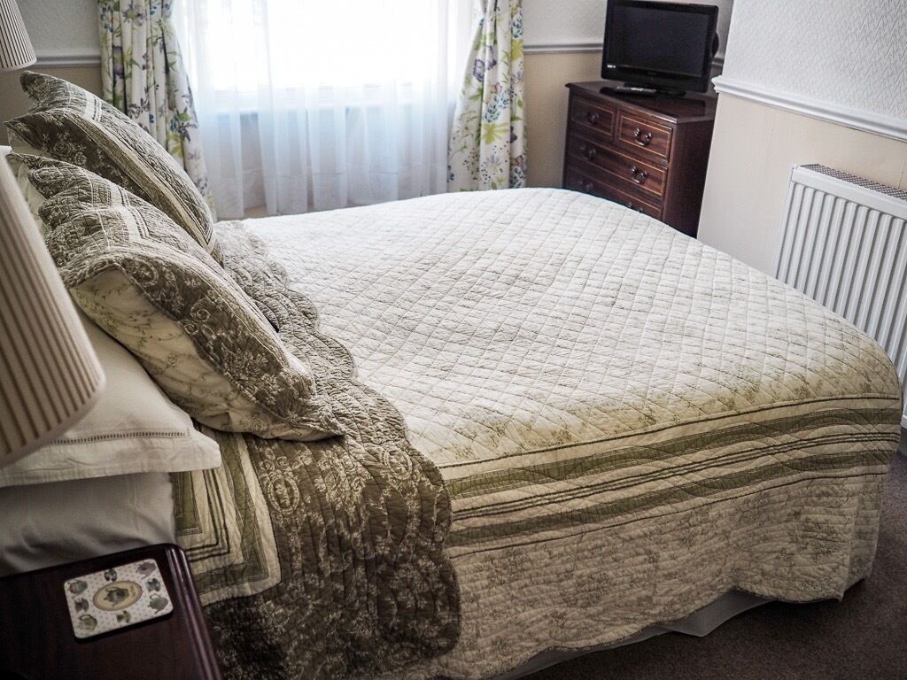 A neatly made bed with a quilted cover, a wooden nightstand with a television, and a radiator in the background.