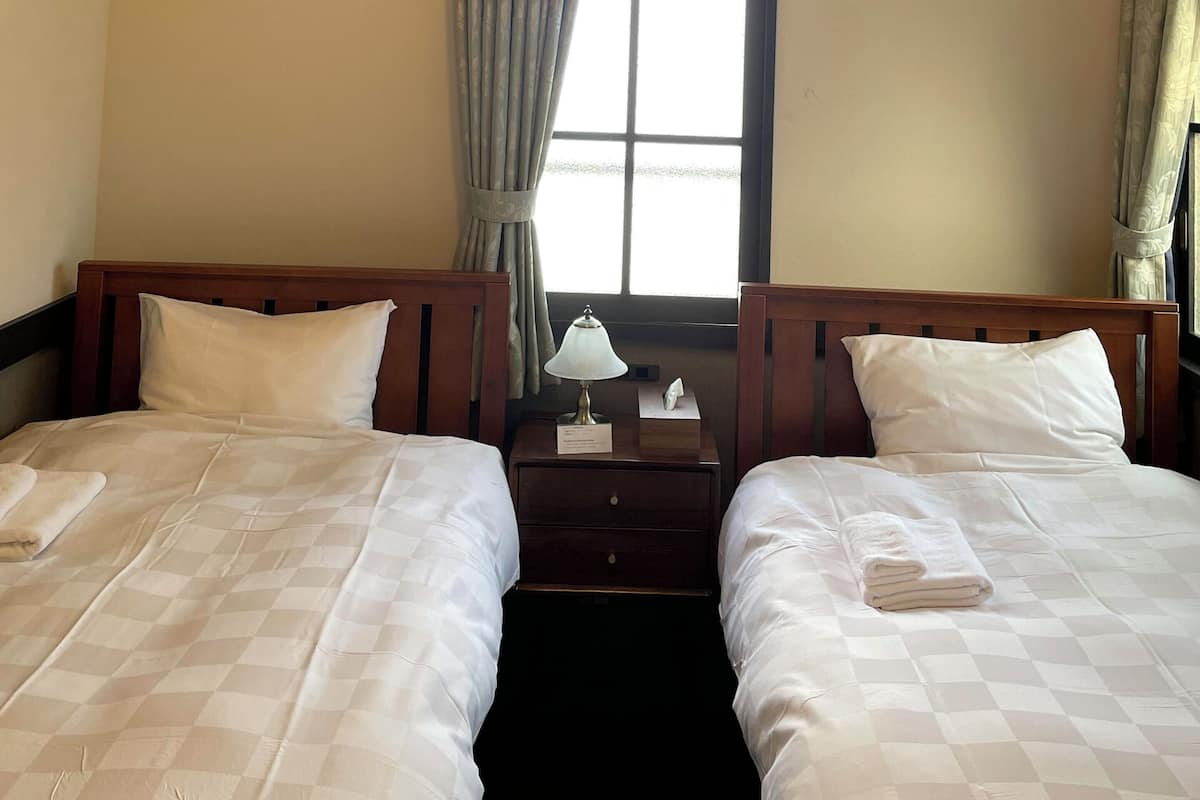A hotel room with two beds, a nightstand, a window with curtains, and a ceiling light fixture.