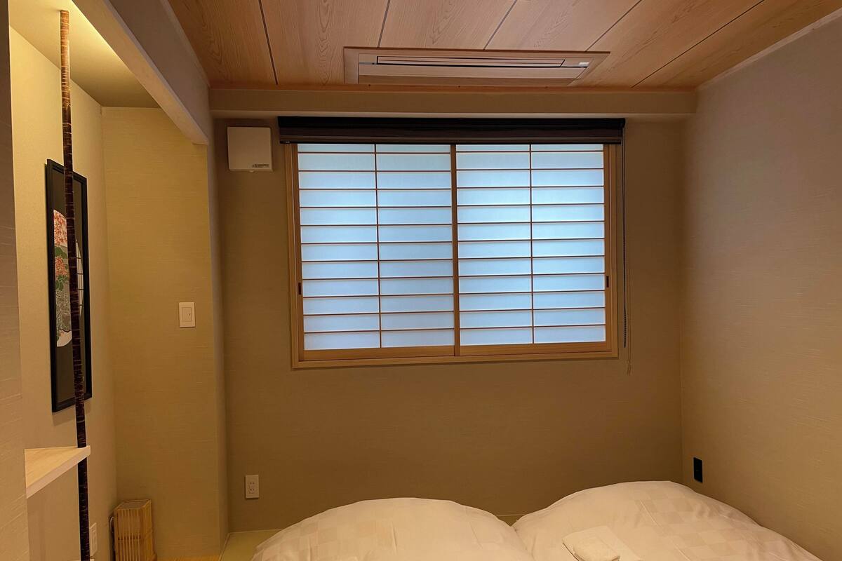 A small, cozy room with a bed, a window with blinds, a shelf, and a radiator.