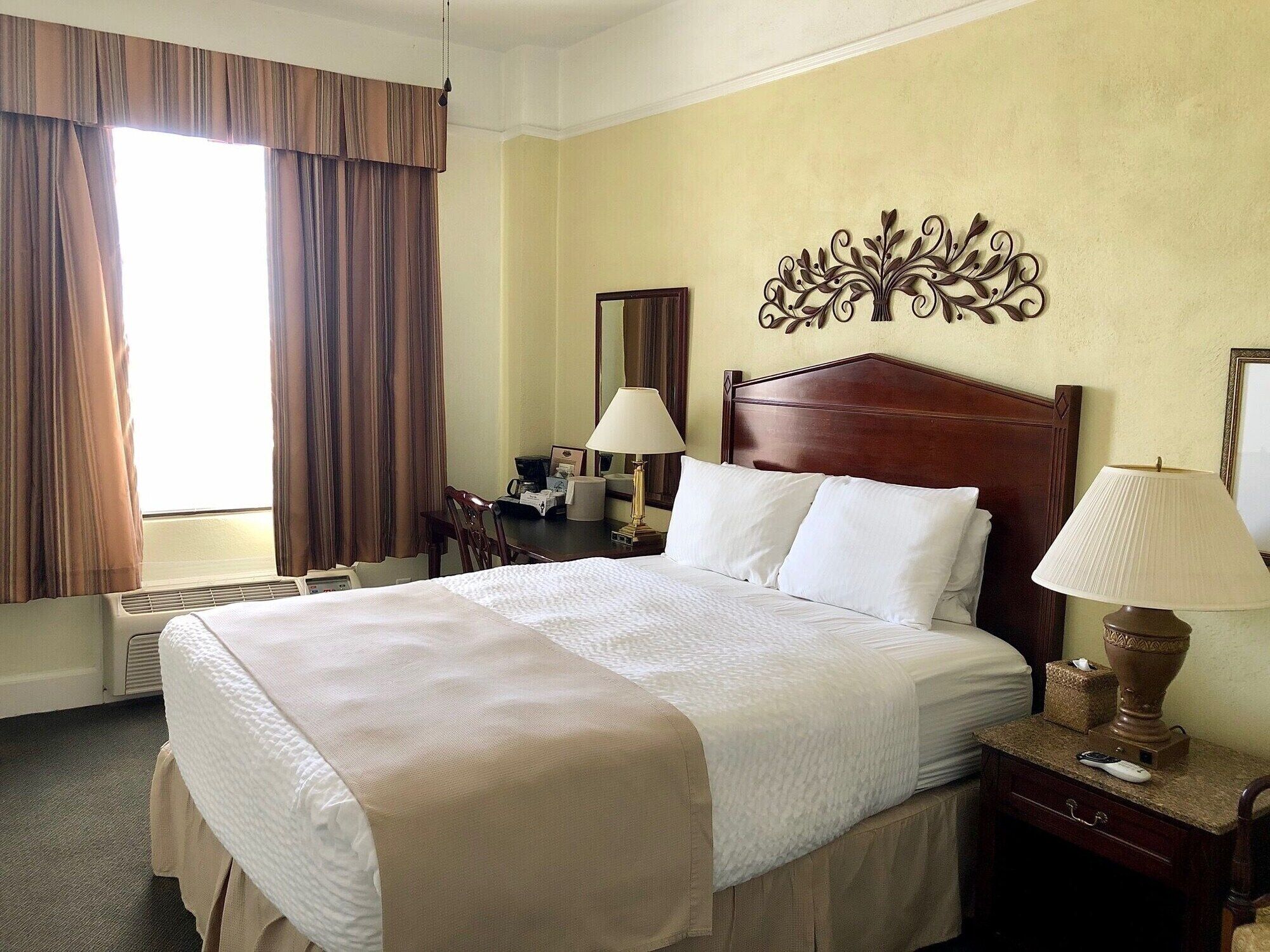 A hotel room with a large bed, two bedside lamps, a desk with a chair, a window with curtains, and a wall decoration.