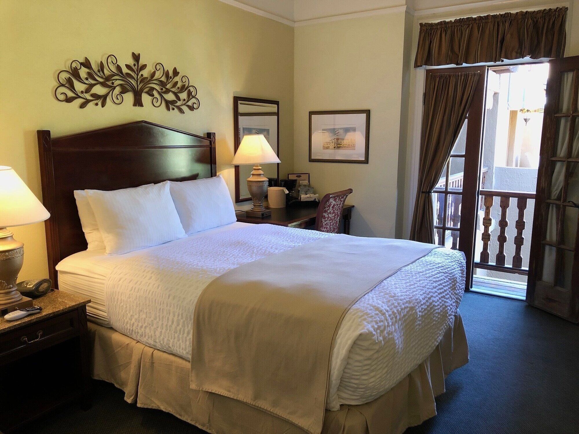 A hotel room with a large bed, two bedside tables, a lamp, a chair, a desk, a window with curtains, and a decorative wall piece.