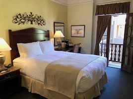 Habitación de hotel con una cama grande, dos mesitas de noche, una lámpara, una silla, un escritorio, una ventana con cortinas y un adorno en la pared.