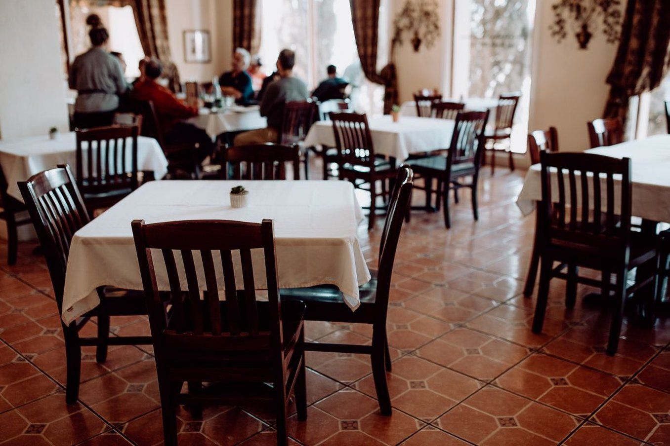 A restaurant interior with multiple tables and chairs, a tiled floor, and a view of diners in the background.