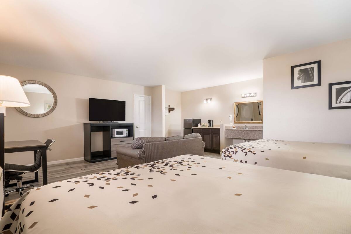 Habitación de hotel con dos camas, un escritorio con silla, televisor, microondas, sofá y baño con espejo, además de cuadros en la pared.
