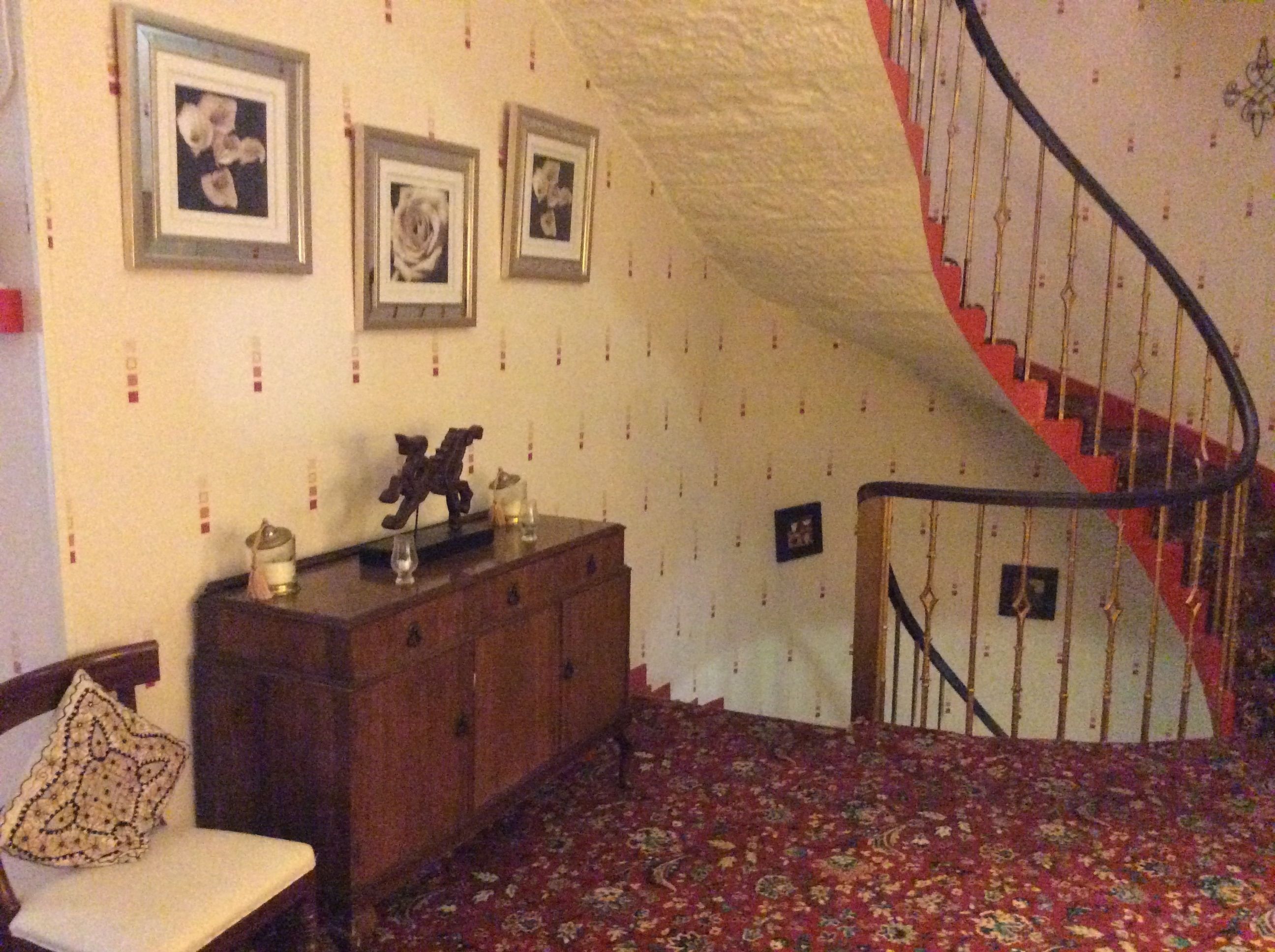 A room with a wooden dresser, a chair, a staircase with a red railing, and three framed pictures on the wall.