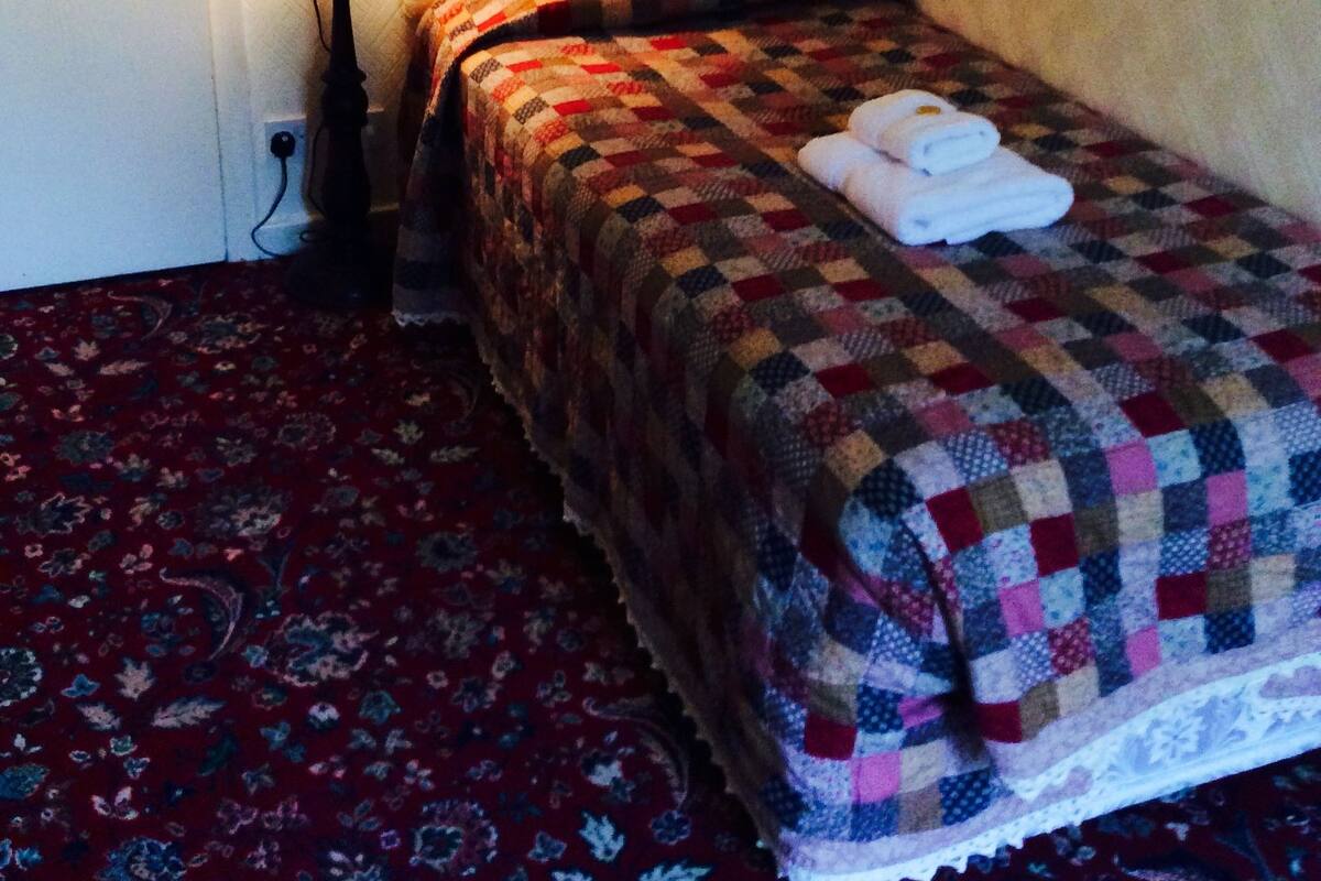 A small, neatly made bed with a checkered blanket and a folded towel on top. A lamp is on a small table beside the bed, and a 'No Smoking' sign is visible on the wall.
