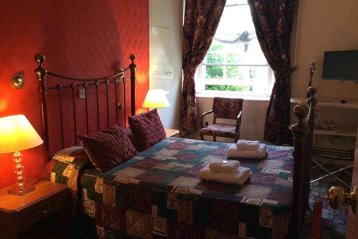 A bedroom with a bed, two pillows, a bedside lamp, a chair, a window with curtains, and a wall-mounted TV.