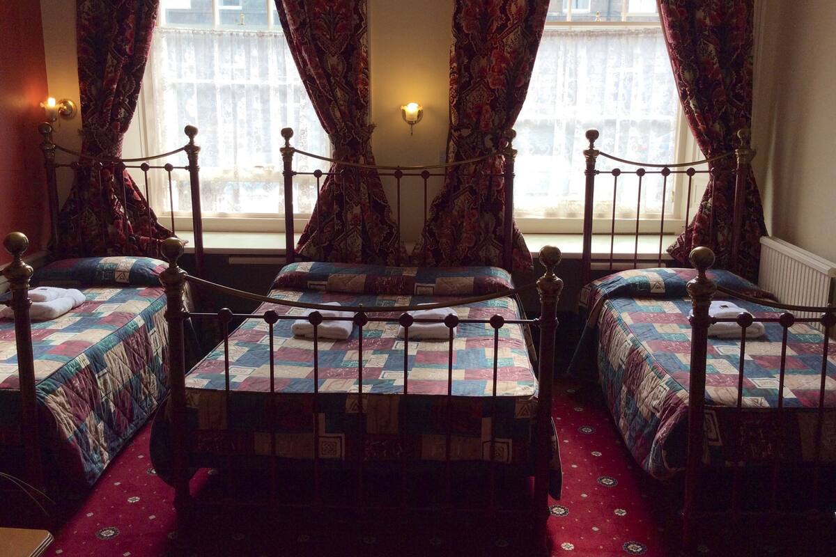 A room with four single beds, each with a metal frame and a quilted bedspread. The room has a red carpet and patterned curtains.