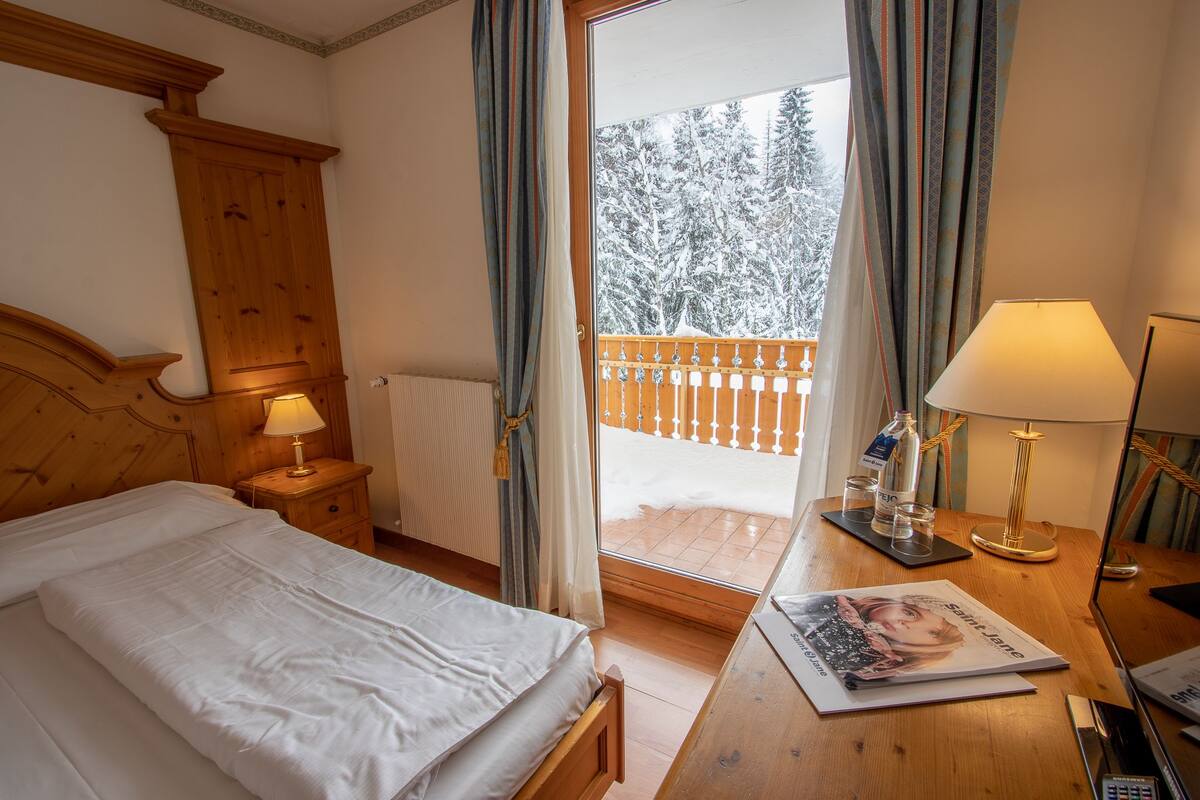 Habitación de hotel con cama, cabecero de madera, mesita de noche con lámpara, mesa con revista y vistas a un balcón nevado.