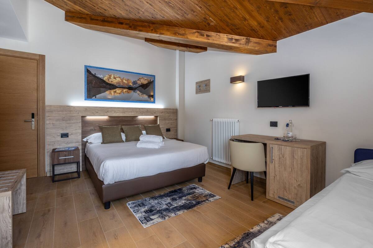 Un dormitorio moderno con techo de madera, una cama con ropa blanca, un escritorio, un televisor y un cuadro en la pared.